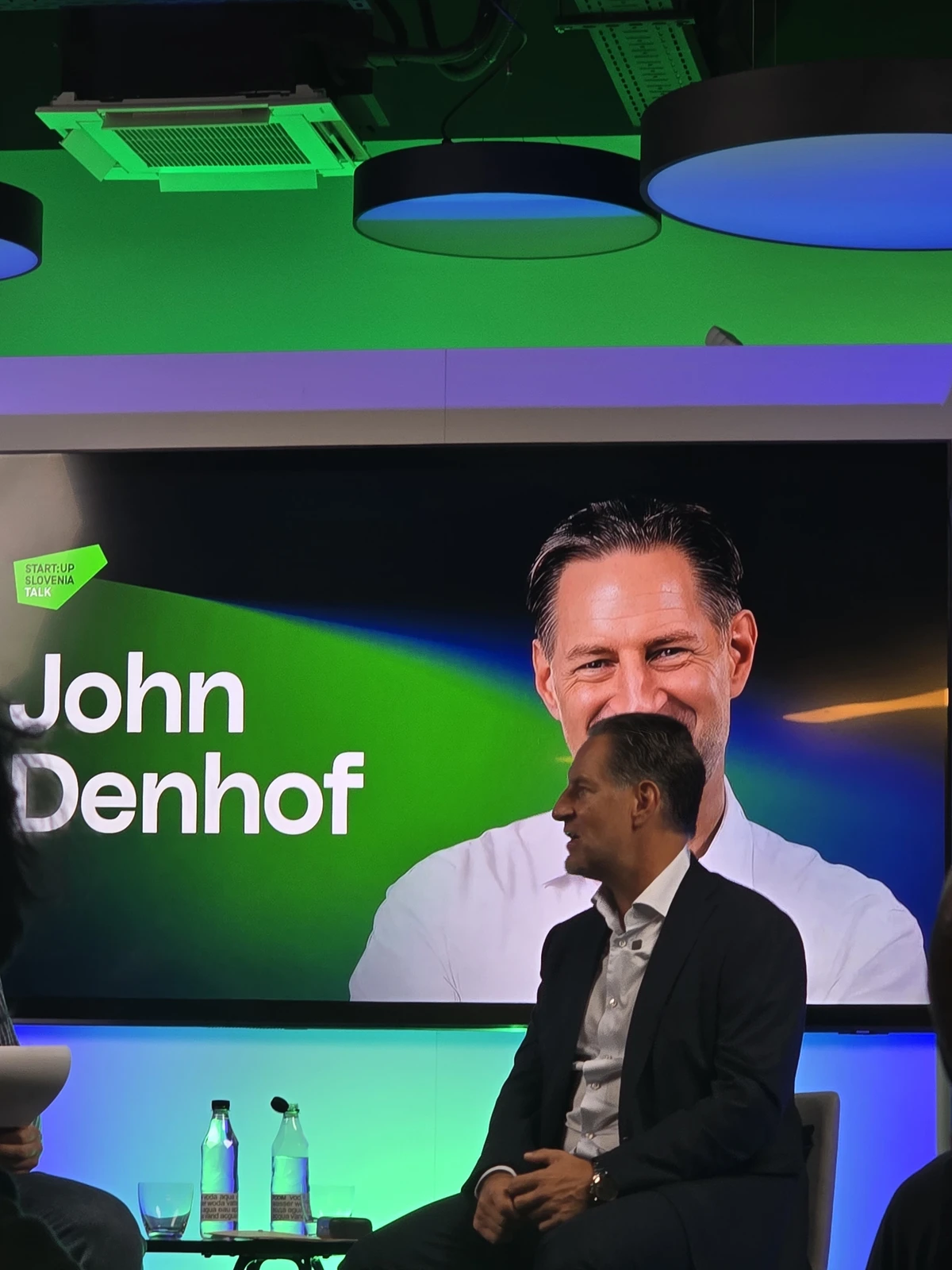 My photo of John Denhof seated on stage in front of his name and portrait at Startup Talk in Maribor
